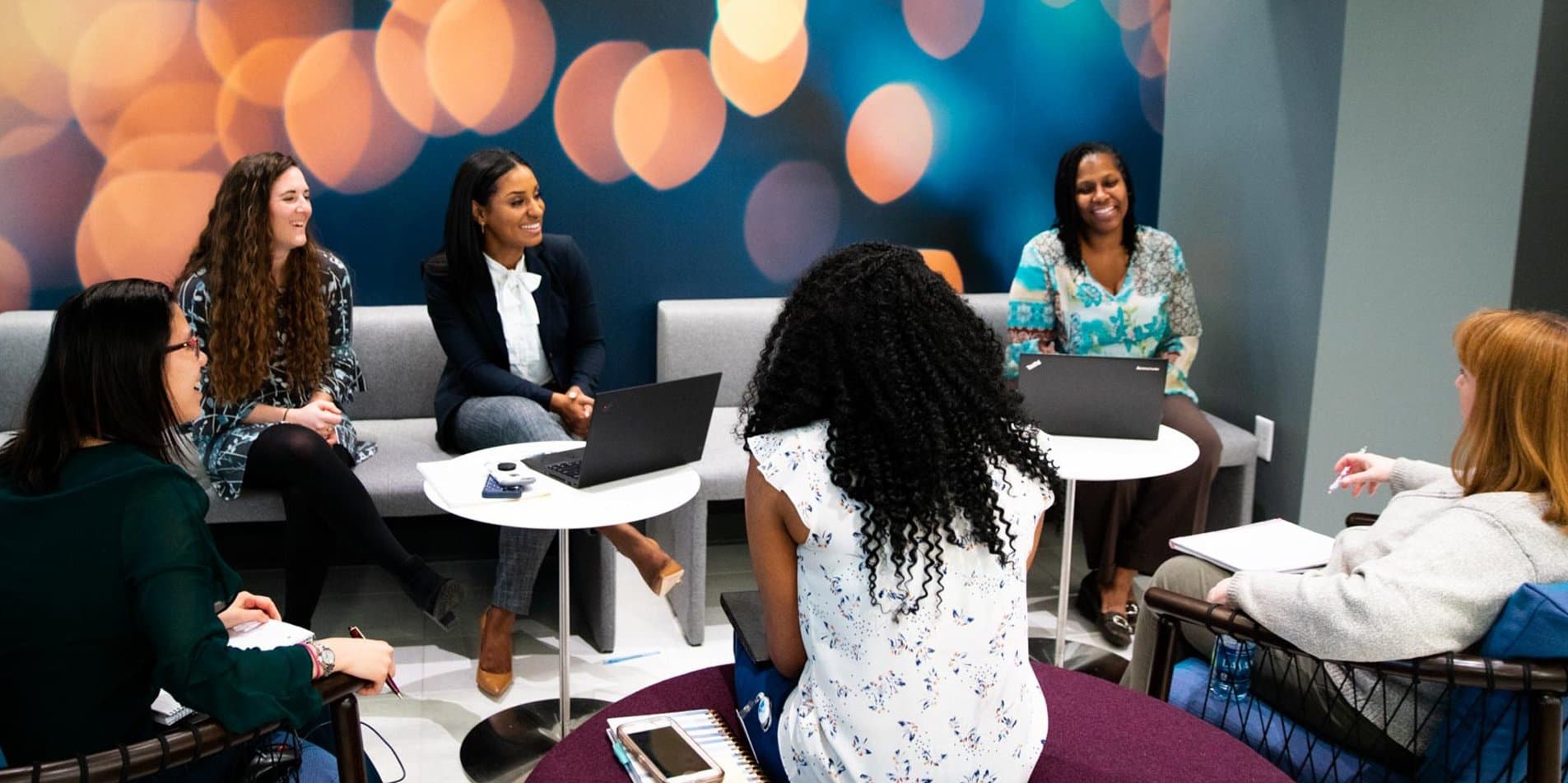 Six people dressed in business casual attire are sitting around an office lounge area on grey sofas and open-backed armchairs. The three people facing the camera are smiling and laughing. The wall behind them is blue with peach circles that look like out-of-focus lights.